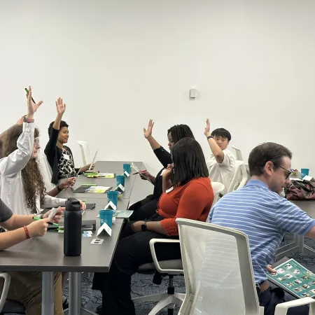 Diverse group in a meeting room with several participants raising hands during a discussion around conference tables.