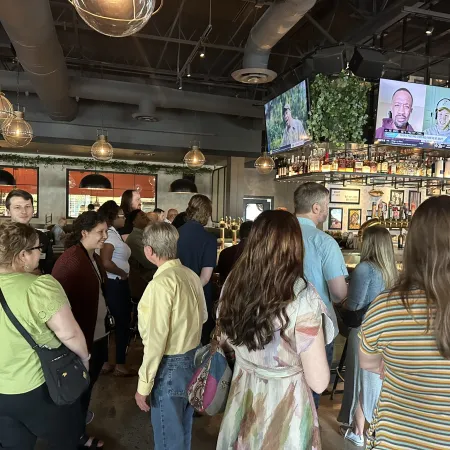 Crowd of people socializing and watching multiple TVs in a modern bar with hanging lights and industrial ceiling.