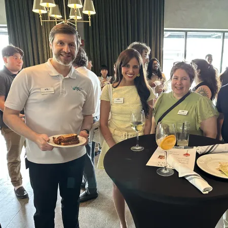 Group of four people smiling at a social event with drinks, food, and name tags in a bright room.