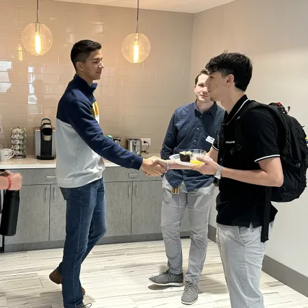 Four young men greeting and conversing in a modern kitchen area with hanging lights and coffee appliances.