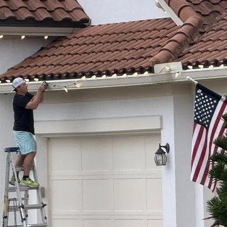 Man on ladder hanging string lights along white house garage roof with American flag nearby under clay tile roof.