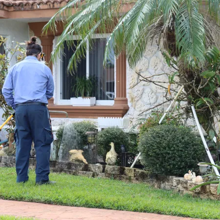 Gardener watering a residential garden with plants, shrubs, and decorative statues near a stone wall.