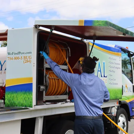 Pest control worker retrieving hose from Royal Pest & Termite truck under clear blue sky on suburban street.