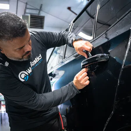 Technician repairing black car door handle in a workshop with focused attention on the task.