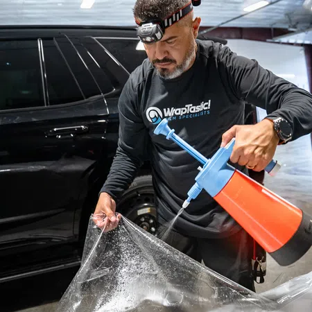 Man wearing headlamp spraying liquid on clear car wrap film in workshop next to black vehicle.