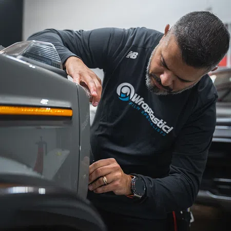Technician carefully applying film wrap to a car's front panel in a professional workshop setting.