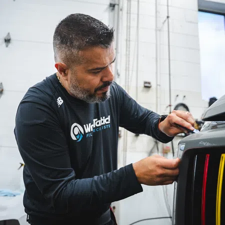 Technician carefully applies detailing work to a black vehicle front in a brightly lit auto workshop.