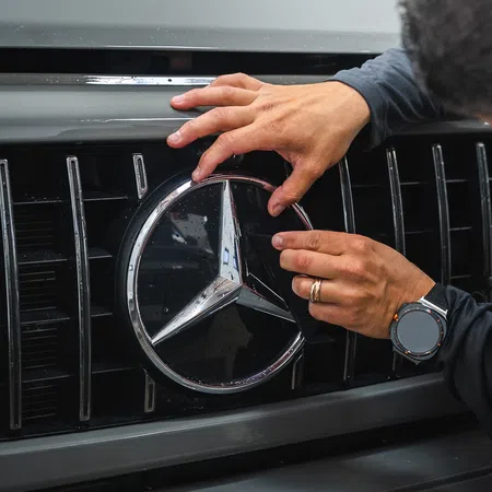 Person cleaning or detailing Mercedes Benz logo on front grille of a gray vehicle with a cloth.