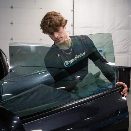 Young man wearing a Watastic! shirt installs window tint film on a black car door in a garage.