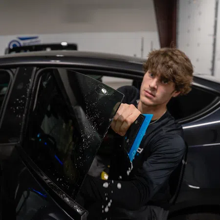Young man installs tinted film on black car window using a blue squeegee tool in a garage.