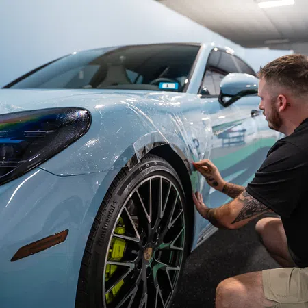 Man applying protective film to front side of a light blue sports car inside a garage.