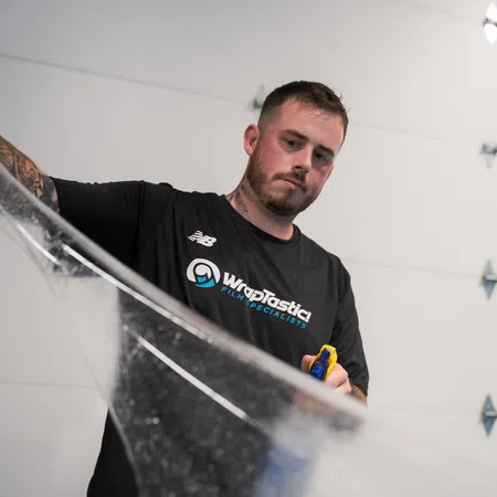 Man wearing a Wraptastic shirt applying protective film using a tool in a garage setting.