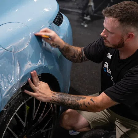 Man with tattoos applying protective film to light blue sports car wheel arch indoors.