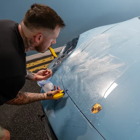 Man applying protective film on blue Porsche hood using spray bottle and yellow squeegee indoors.