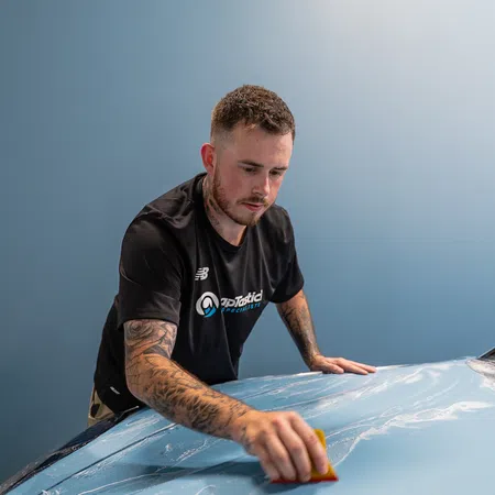 Man with tattoos carefully applying protective film on a car hood using a squeegee in a workshop.