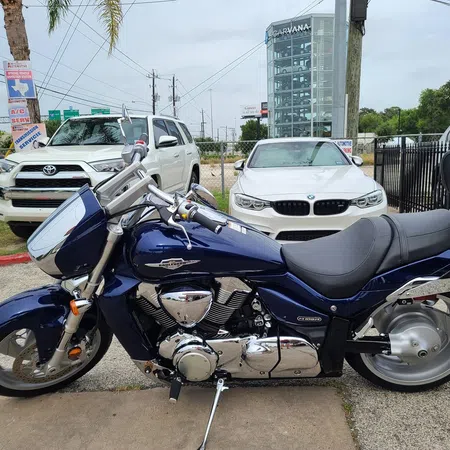 Blue Suzuki Boulevard motorcycle parked next to white Toyota SUV and BMW sedan in urban setting.