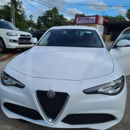 Front view of a white Alfa Romeo car parked outside an automotive service center under a blue sky.