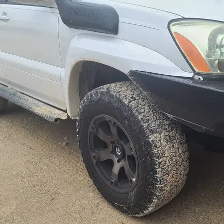 Close-up of a white off-road SUV front tire with mud and black custom rims on a dirt road.