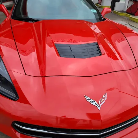 Shiny red Chevrolet Corvette hood with black vent and Chevrolet emblem at gas station background.