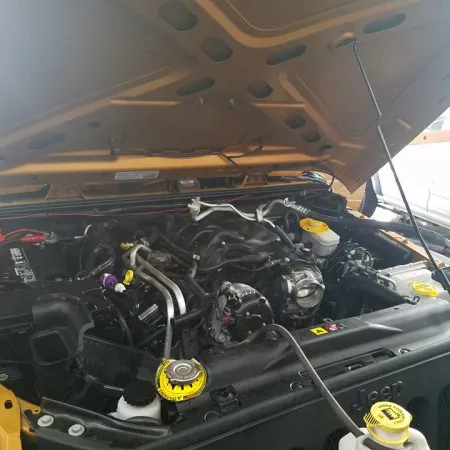 Open hood of yellow Jeep showing detailed engine components and fluid reservoirs in a garage setting.
