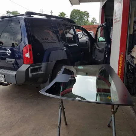Blue Nissan SUV at auto repair shop with removed windshield placed on a stand ready for replacement.