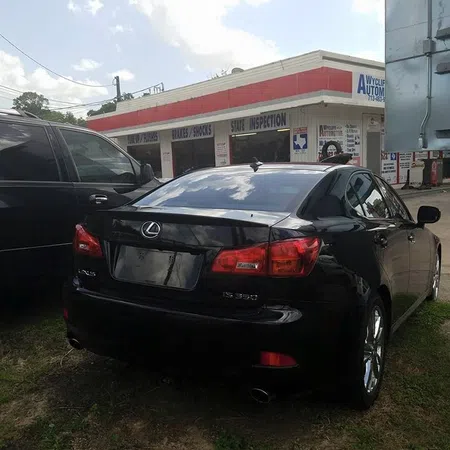 Black Lexus IS 350 parked near a repair shop with signs for brakes, shocks, and state inspection under a cloudy sky.