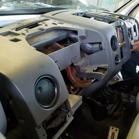 Dashboard of a vehicle removed for repair showing internal air vents and steering components with mechanic working