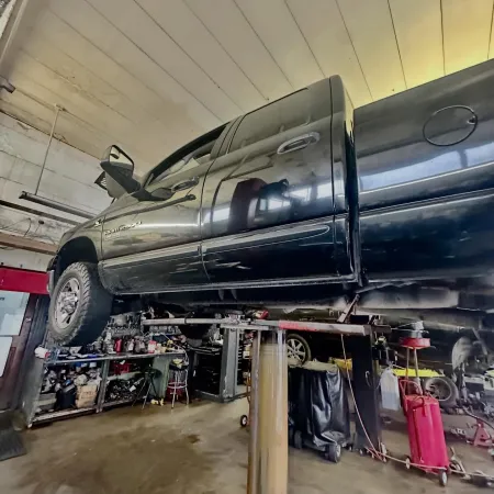 Black pickup truck lifted on a hydraulic car lift inside a cluttered auto repair shop.