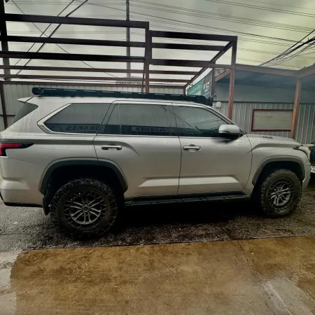Silver off-road SUV parked on wet concrete under metal frame structure with cloudy sky.