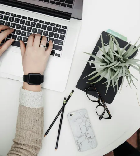 Person typing on laptop at a white round table with plant, glasses, phone, and pencils nearby.