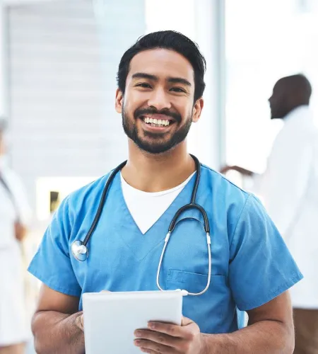 Smiling male nurse in blue scrubs with stethoscope holding a tablet in a bright hospital setting.