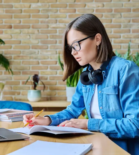 Young woman in glasses and denim jacket writing in notebook with laptop and headphones on wooden desk