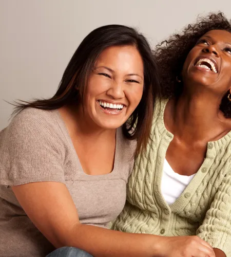 Three diverse women sitting closely and laughing joyfully on a beige couch in casual attire.