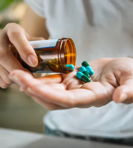 Person pouring blue and green capsules from a brown pill bottle into an open hand indoors.