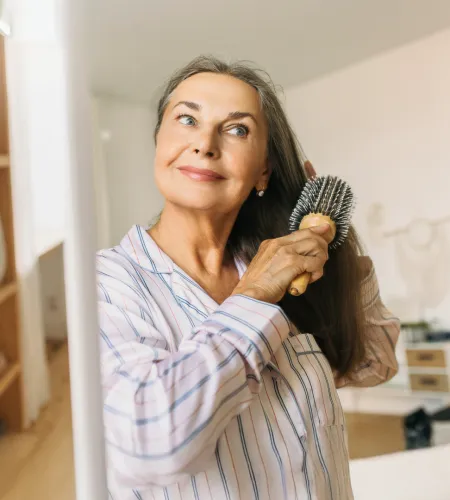 Smiling middle-aged woman brushing her long hair in front of a mirror wearing striped pajamas.