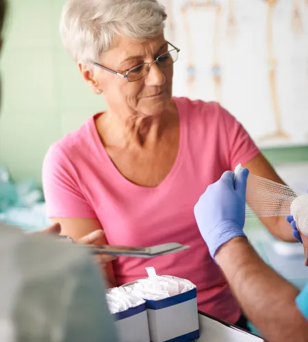 Medical professional wrapping bandage on elderly woman's hand in a clinic setting with anatomical chart in background