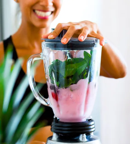 Smiling woman making a green smoothie with fresh spinach and pink liquid in a blender at home