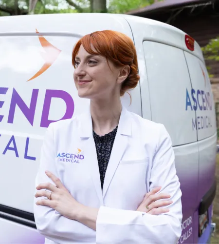 Confident female medical professional in white coat stands with crossed arms beside Ascend Medical van outdoors.