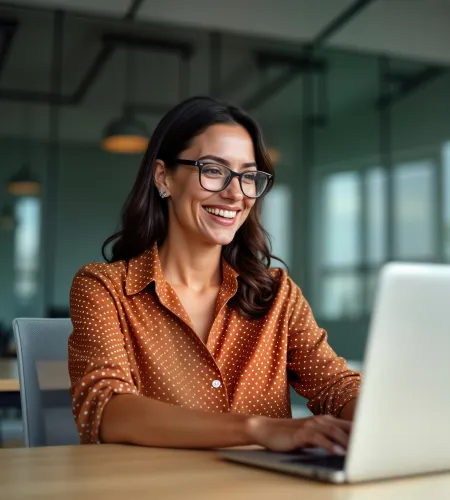 Smiling young woman in glasses working on laptop in modern office with large windows and plants.