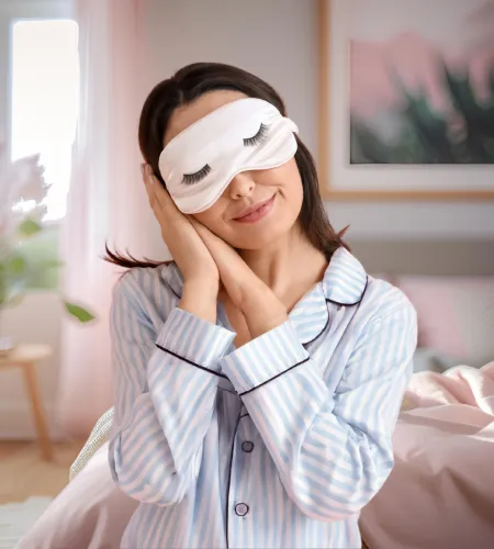 Woman in striped pajamas wearing a sleep mask with eyelashes, resting her head on hands in a cozy bedroom.