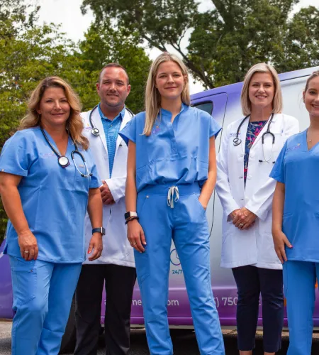 Group of five medical professionals in blue scrubs and white coats standing outside by a medical vehicle.
