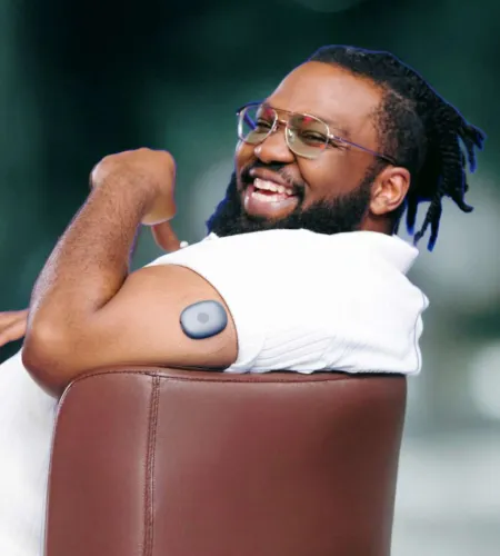 Smiling man with glasses showing wearable health device on upper arm while sitting on a chair outdoors.