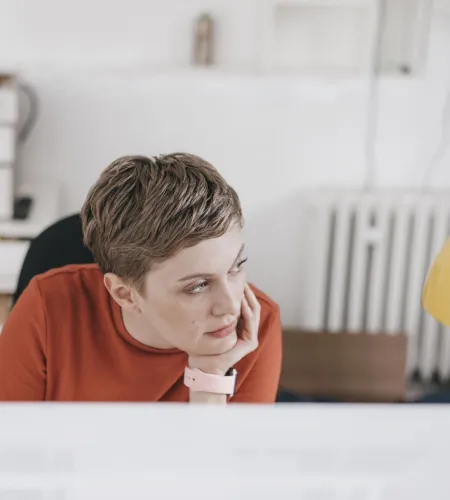 Young woman in red sweater looking thoughtfully away while sitting at desk in a modern office space.