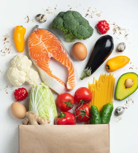 Fresh salmon, vegetables, eggs, avocado, pasta, and rice spilling from a paper grocery bag on white background