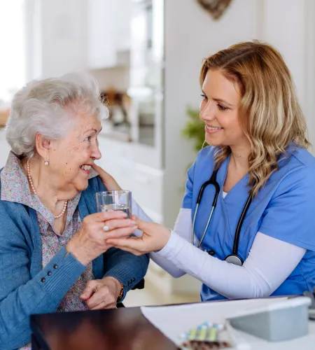 Nurse assisting elderly woman with a glass of water in a bright, cozy living room.