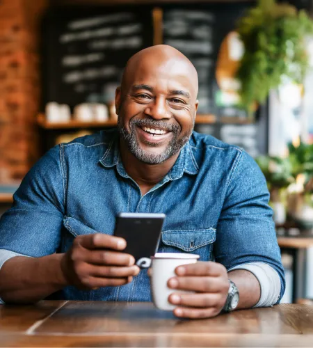 Smiling man in denim shirt sitting at cafe table holding smartphone and coffee cup in cozy setting