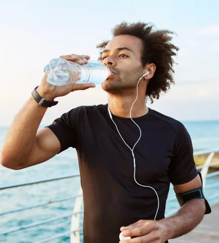 Man in black shirt drinking water while standing by the ocean with earphones and armband on his arm.