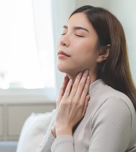 Young woman sitting indoors with eyes closed, gently touching her throat, appearing to soothe or relieve discomfort.