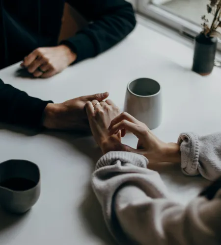 Couple holding hands across a white table near coffee cups and a window for intimate conversation.