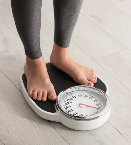 Person standing barefoot on a white analog bathroom scale on a light wooden floor.